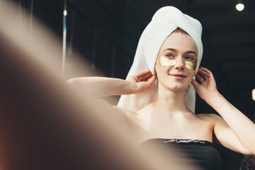 Caucasian woman is smiling in the mirror applying golden hydrogel eye patches and wearing a towel