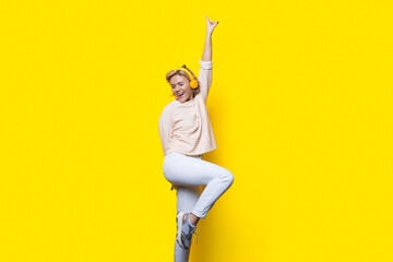 Sexy blonde lady is smiling and dancing on a yellow studio wall wearing headphones