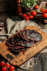 Delicious smoked sausage on a dark wooden board with tomatoes and spices. Delicious handmade snack made from natural ingredients. Pieces of dry pork, beef, chicken snacks. Meat products. Close-up.