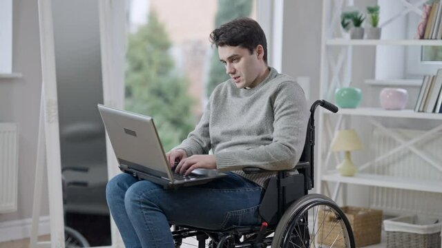 Portrait of positive handsome young paraplegic man in wheelchair talking messaging online using laptop. Brunette Caucasian paralyzed disabled guy indoors at home. Handicap and lifestyle