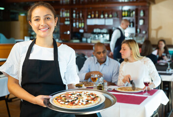 Hospitable latin american waitress holding plate with appetizing pizza, meeting guests in cozy pizzeria