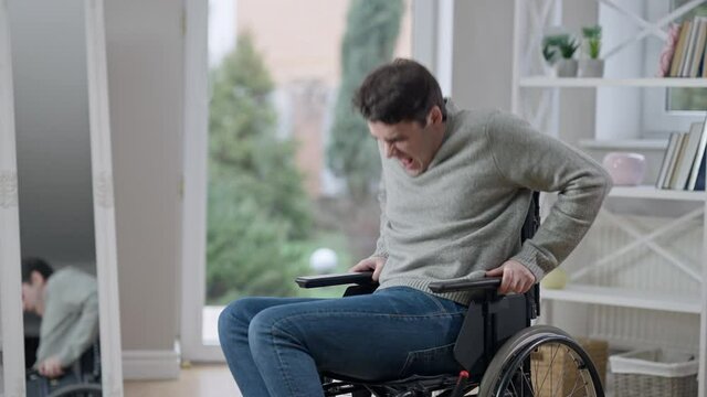 Handsome concentrated young paraplegic man with paralyzed legs trying to stand up of wheelchair indoors. Portrait of confident motivated Caucasian invalid recovering after trauma. Effort and medicine