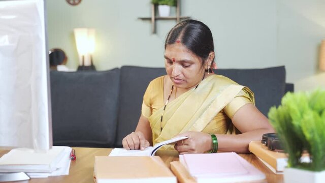 Indian woman busy reading or searching on book by pointing finger on sentence while working from home.