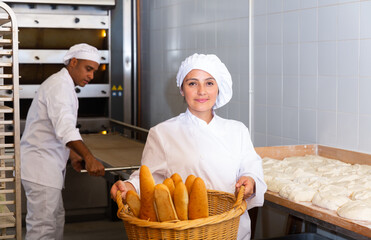 Female baker holding basket of hot baguettes in the kitchen of the bakery