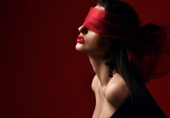 Profile of excited woman with naked shoulders and breast holds and eyes covered with red scarf, blindfold over dark background with copy space. Fashion, vogue, sexy stylish look for woman concept - 424345527