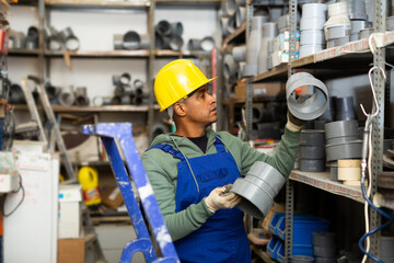 Salesman in overalls with plumbing fittings at a hardware store warehouse - obrazy, fototapety, plakaty