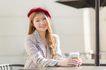Asian beautiful woman who wears suit and red cap with bronze hair sits on chair in coffee shop while holds coffee cup in her hand on a sunny morning.