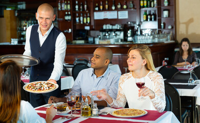 Fototapeta na wymiar Polite waiter bringing pizza to friends in cozy restaurant