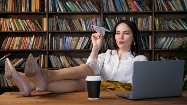 Joyful secretary in white blouse and yellow skirt plays with paper plane putting legs on table with grey laptop and coffee cup in office