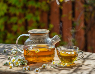 Herbal chamomile tea and chamomile flowers near teapot and tea glass on wooden table. Countryside...