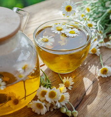 Herbal chamomile tea and chamomile flowers near teapot and tea glass on wooden table. Countryside background.