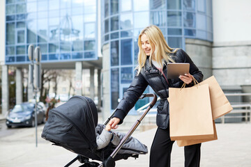 Mother with baby carriage holding paper bag and tablet on city street in spring. Shopping, parenthood and lifestyle concept.