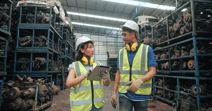 Two workers technical young Asian female and male engineers with safety suit helmet discussion together on process of work by digital tablet in used auto parts factory industrial.
