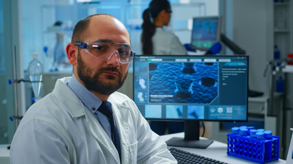 Portrait of exhausted scientist man looking at camera sitting in front of computer with DNA scan image. Chemist examining virus evolution using high tech and chemistry tools for scientific research