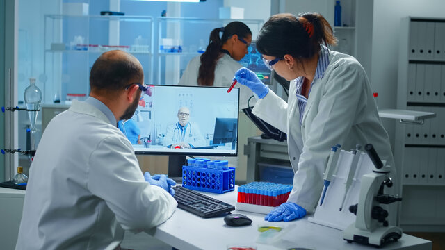Lab technicians talking on video call with professional chemist doctor explaning vaccine reactions, woman nurse showing blood sample on web cam, during virtual meeting in medical research laboratory