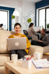 Fototapeta premium Young woman entrepreneur sitting on couch in middle of business start up office looking at camera smiling holding laptop. Multiethnic coworkers talking about startup financial company in modern office