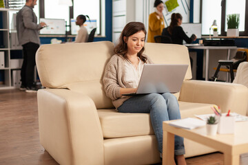 Obraz premium Focused manager reading tasks on laptop and typing sitting on couch in busy start up office while diverse team analyzes statistics data in background. Multiethnic team talking about company project