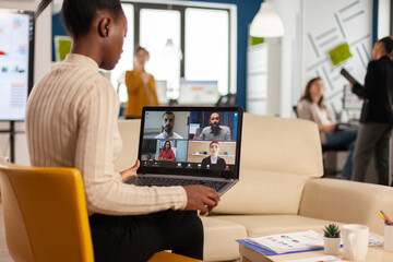 Fototapeta na wymiar African manager woman discussing with remote colleagues using video call holding laptop sitting on couch in business modern office. Diverse coworkers planning new financial project in modern company
