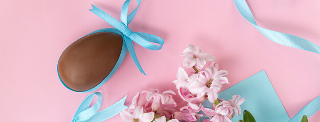 banner with Easter background with chocolate egg with blue bow and pink hyacinth flowers, with blue ribbon on pastel pink and blue colors. Happy Easter concept.