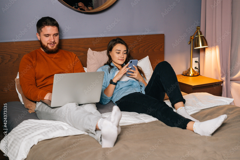 Wall mural Cheerful loving young couple lying in bed with their devices at evening. Handsome bearded man working typing on laptop and pretty young woman using mobile phone. Concept of leisure activity at home.