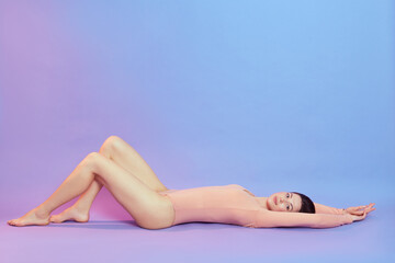 Obraz premium Beautiful young woman with long legs wearing beige bodysuit lying on floor and looking up, female with perfect body raising legs, posing isolated over blue background with pink neon light.