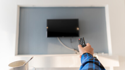 A young man with a remote control and a coffee mug is watching TV in the room.