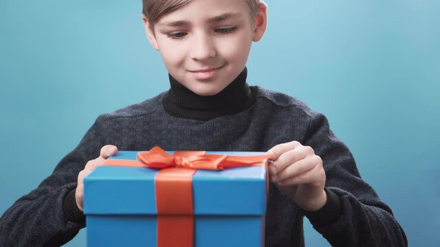 Happy blonde boy with blue eyes opens a gift box on a blue background and is surprised, light is shining from the box. Celebration concept, birthday, new year, christmas.