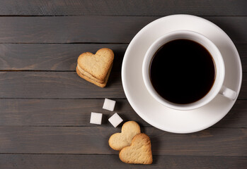black coffee in a white cup on a white saucer with heart-shaped cookies and sugar cubes on a brown wooden background horizontal photo top view