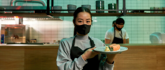 Portrait of young waitress wearing protective mask looking at camera and holding a plate with sushi rolls. Japanese cuisine in restaurant.