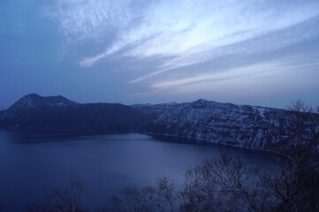 薄い雲の広がる夜明けの空と山間の湖。日本の北海道の摩周湖。