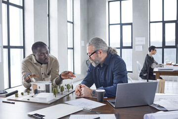 Two architects sitting at the table with model of new building and discussing new project at office