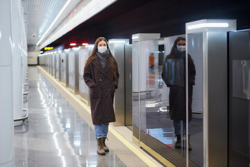 A full-length photo of a woman in a medical face mask to avoid the spread of coronavirus who is standing on the subway platform. A girl in a mask is keeping social distance.