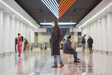 A full-length photo of a woman in a face mask to avoid the spread of COVID who is walking while waiting for a train in the center of a metro station. Girl in a surgical mask is keeping social distance