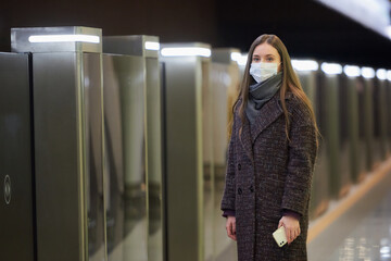 A lady in a medical face mask to avoid the spread of coronavirus is waiting for a train and holding a cellphone at the subway station. A girl in a surgical mask is keeping social distance in the metro