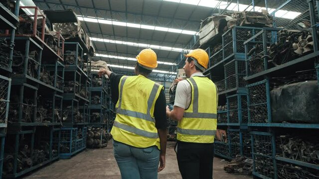 Two workers technical with safety suit helmet walking around car industry checking stock of used auto parts by digital tablet in factory industrial