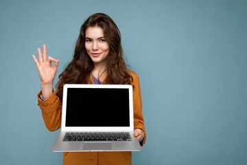 Photo of Beautiful satisfied happy young woman holding computer laptop looking at camera wearing yellow jacket showing ok gesture isolated over blue wall background