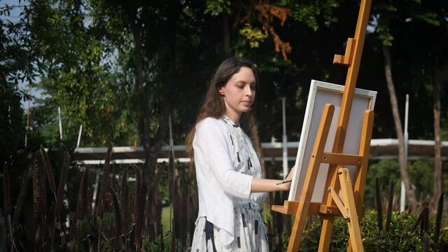 Girl artist in a white dress at work in the flower garden. Doing oil painting on canvas. With loose hair, concentrated, focused, hard-working and professional.