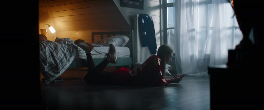 Attractive Caucasian teenager girl lying on the floor, using phone, chatting with friends in her attic bedroom. Shot with 2x anamorphic lens