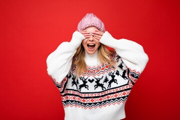 Photo of young positive happy beautiful blonde lady with sincere emotions wearing pink knitted hat and winter pullover isolated over red background with empty space and covering eyes with hands