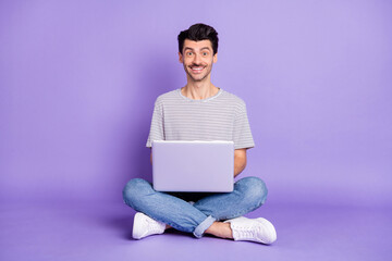 Photo of man sit floor hold pc toothy beaming smile wear black t-shirt jeans sneakers isolated purple background