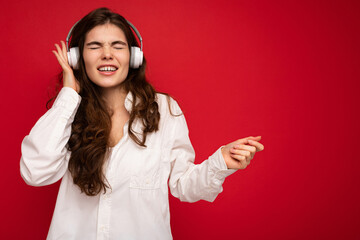 Attractive emotional young brunette female person wearing white shirt and optical glasses isolated over red background wearing white wireless bluetooth earphones listening to music with sincere