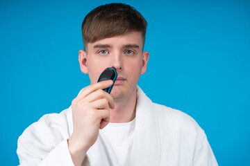 Beauty portrait of handsome guy, young man in white bath robe using electric razor or shaver to remove, plucking excess hair from nose. Hair, skin care. Removing hair with trimmer. Personal hygiene.