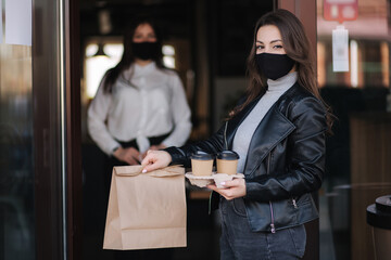 Attractive female customer in face mask standing outdoors by cafe and take away food and coffee. Background of cashier in mask waiting for next customer. New norm during quarantine. Covid19