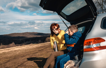 Mother and son take break from driving sitting by the car while drinking tea in nature.
