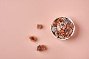 Brown caramelized lump cane sugar cubes in a bowl on pink powdered background, top view, copy space