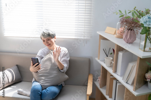 Elderly woman using smartphone video conference with grandchild while lying on sofa in living room at home. Enjoying time lifestyle senior family at home concept. Portrait looking at camera