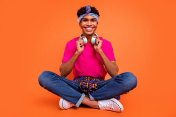 Full length body size photo of happy young man wearing wireless headphones smiling sitting down isolated vibrant orange color background