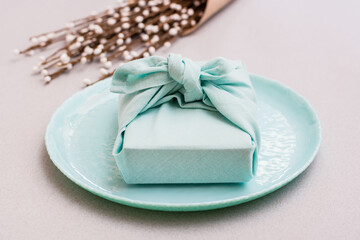 Eco-friendly gift - a box wrapped in cloth on a plate and a bouquet of pussy willow on a gray background. Minimalism.