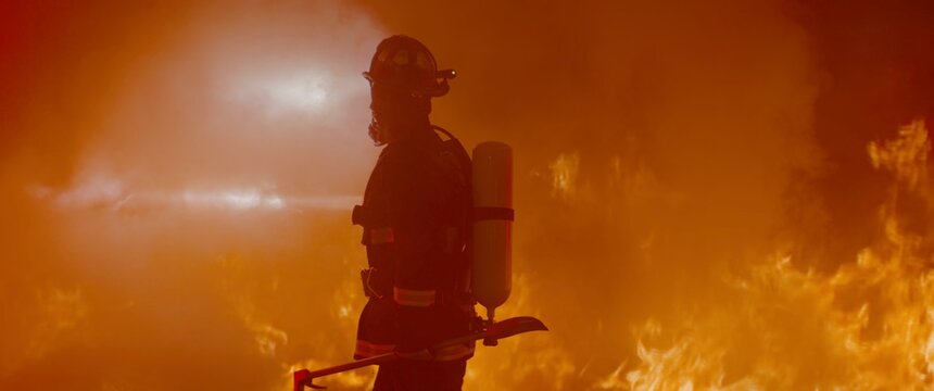 Dramatic silhouette of American firefighter in full gear exploring the huge fire zone