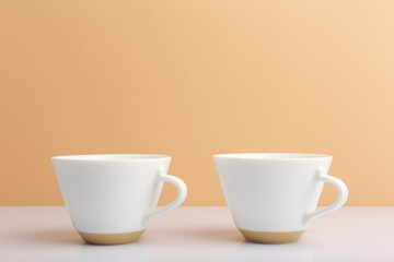 Close up of two white ceramic coffee cups on white table against beige background